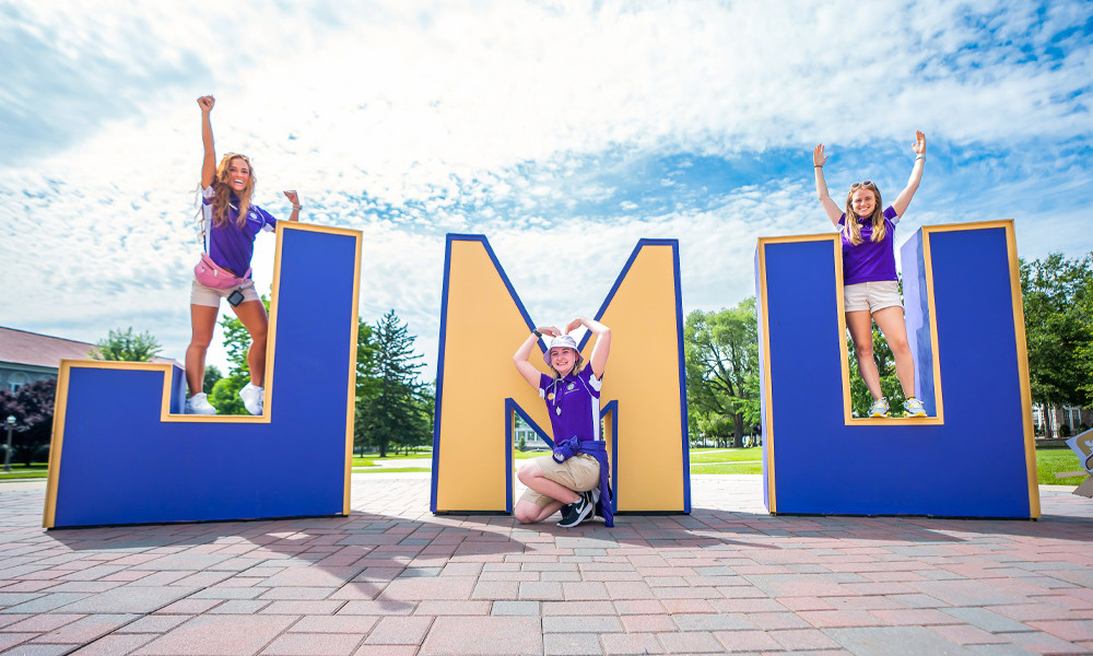 Three women stand on over-sized JMU letters on the quad.