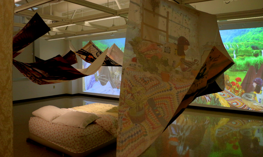 Art installation - bed in the middle of a room with a projection on one wall and hanging prints