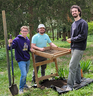 Three students standing around a sifting table in a grassy yard.