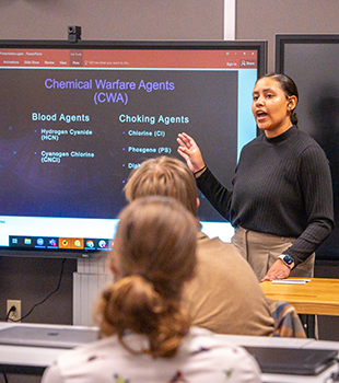 A young woman gestures to a slide of her presentation on a projector screen, speaking towards a seated crowd.