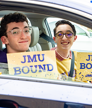 Two young students sit in a car with the window rolled down, holding up signs that read "JMU BOUND".