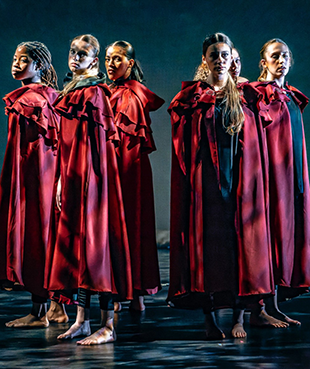 A group of student performers standing in a circle wearing red cloaks.