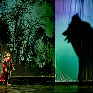 A student performer wears a red cloak and looks up towards a huge shadow of a wolf on a stage screen.