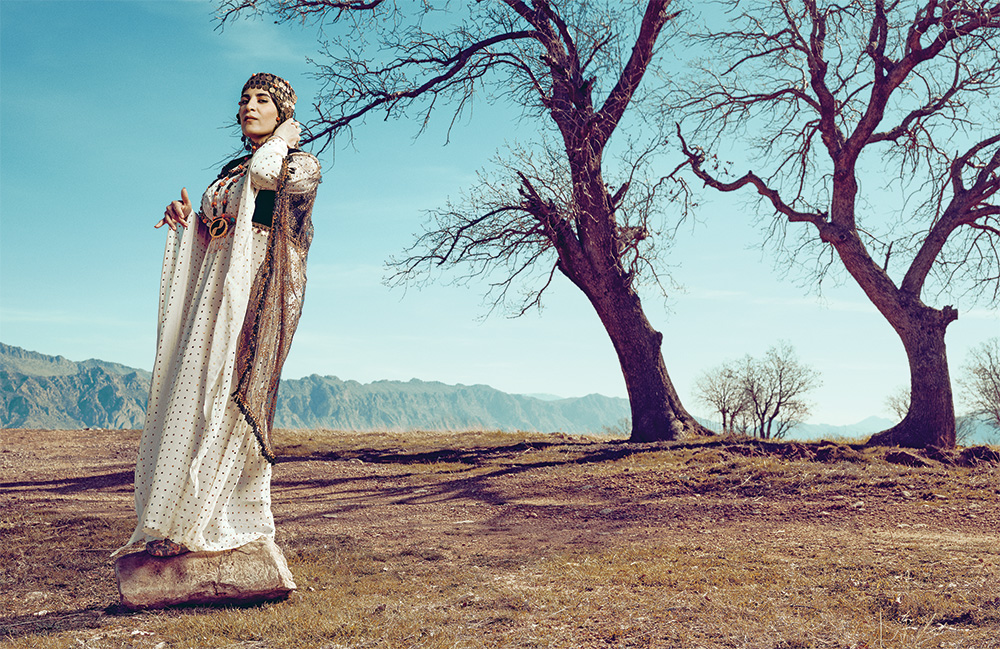 A woman wearing traditional clothing stands on a rock by two trees with no leaves, a mountain range in the background stretches into the distance.