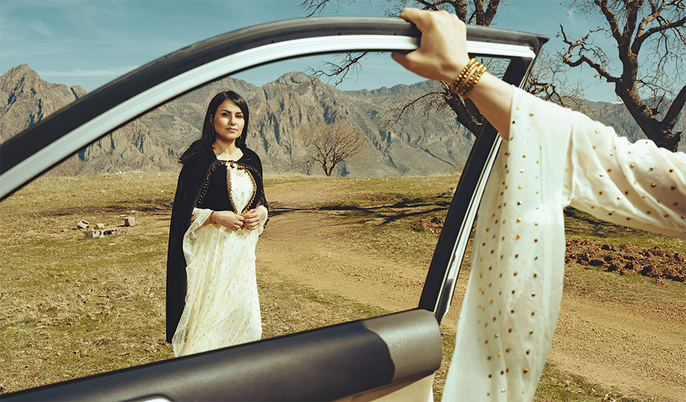 A woman in traditional clothing is framed by a car door window as a hand with a bracelet grips the top of the car door, in the background on rocky mountains.
