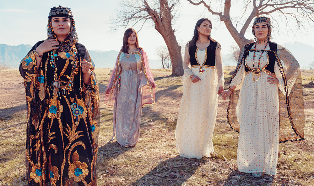 Four women wearing traditional clothing stand side-by-side, they each cast shadows; in the background are two leafless trees, a mountain range is barely visible further away.