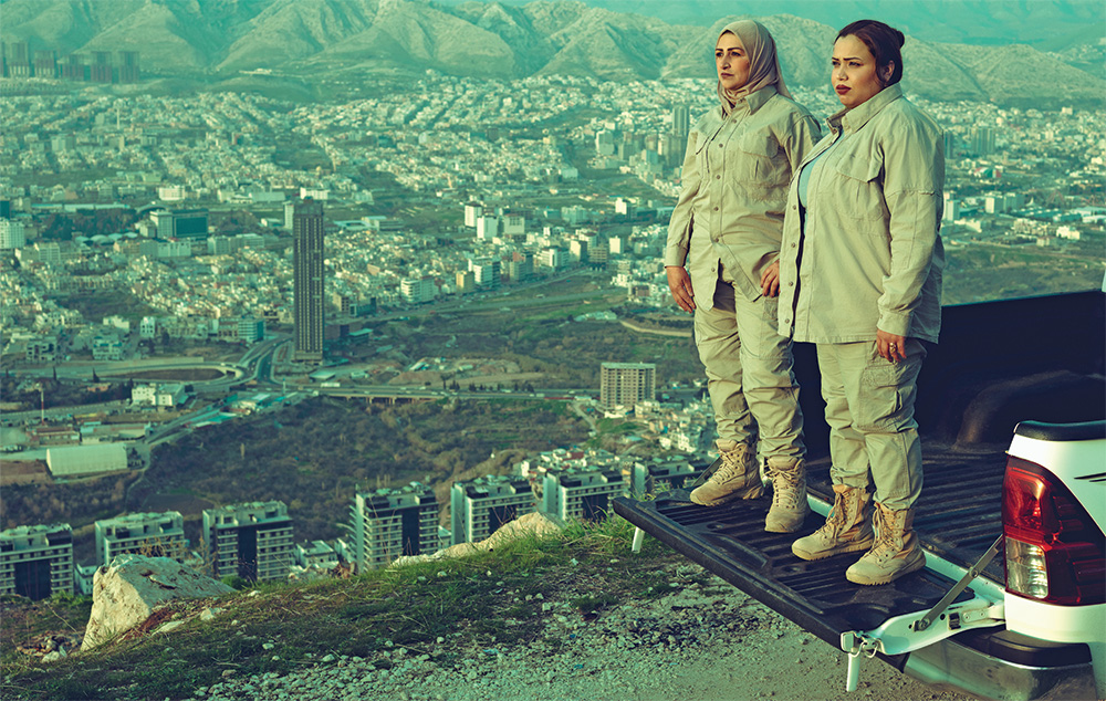 Two women in tan fatigues stand on the tailgate of a pickup truck that is parked overlooking a city with tall apartment surrounded by mountains.