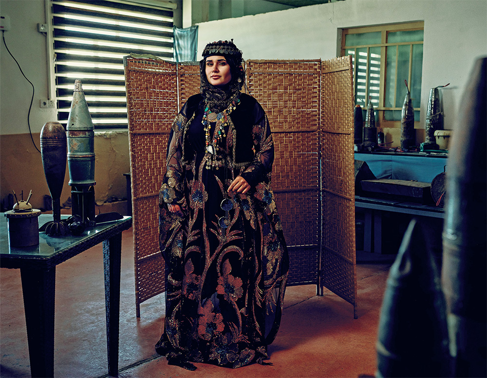 A woman in black traditional clothing with gold and navy embroidery stands in a room with multiple items of landmines and explosive ordnance stacked on surrounding tables.