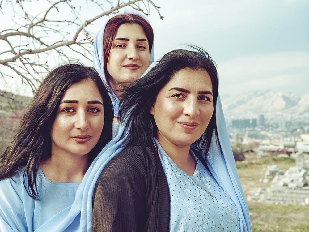 Three women wearing blue dresses and scarves, smile and pose closely together for a picture with a city and mountains in the background.