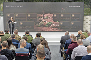 Audience members watch a  woman stands at a podium at the front of a room in front of a projection screen showing a person in military clothing laying on his stomach.