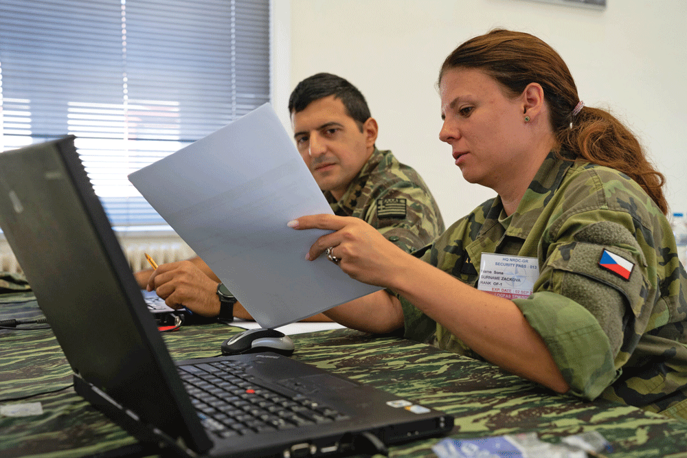 Two people in military clothing sit in front of a laptop computer looking a a piece of paper one of them is holding.