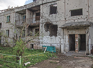 A partially ruined building with battle damage including bullet holes, missing doors and windows. There is grass and a yellow flowers beneath a hole in the wall on the first floor.