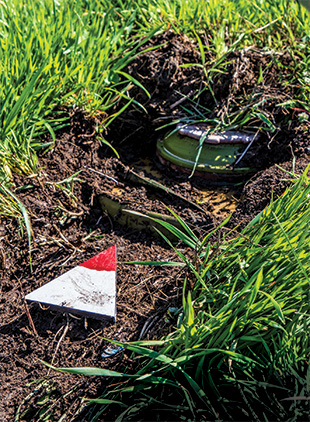 A wooden triangle with a painted red tip pointing toward a partially buried green, metal object surrounded by thick grass.