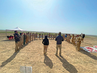 A group of people wearing tan fatigues stand in a crescent shape as two people address the group.