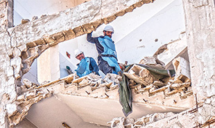 Two men wearing blue personal protective vests and white helmets walk down stairs of a partially destroyed building.