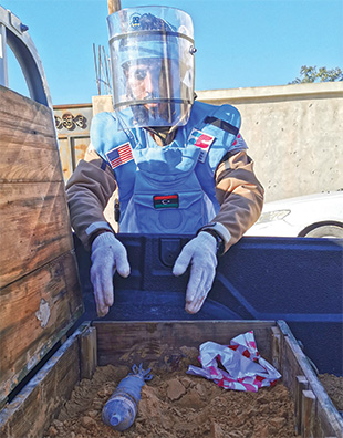 A man wearing blue personal protective equipment and a polycarbonate visor reaches into a truck bed to grab a munition from a crate full of dirt.