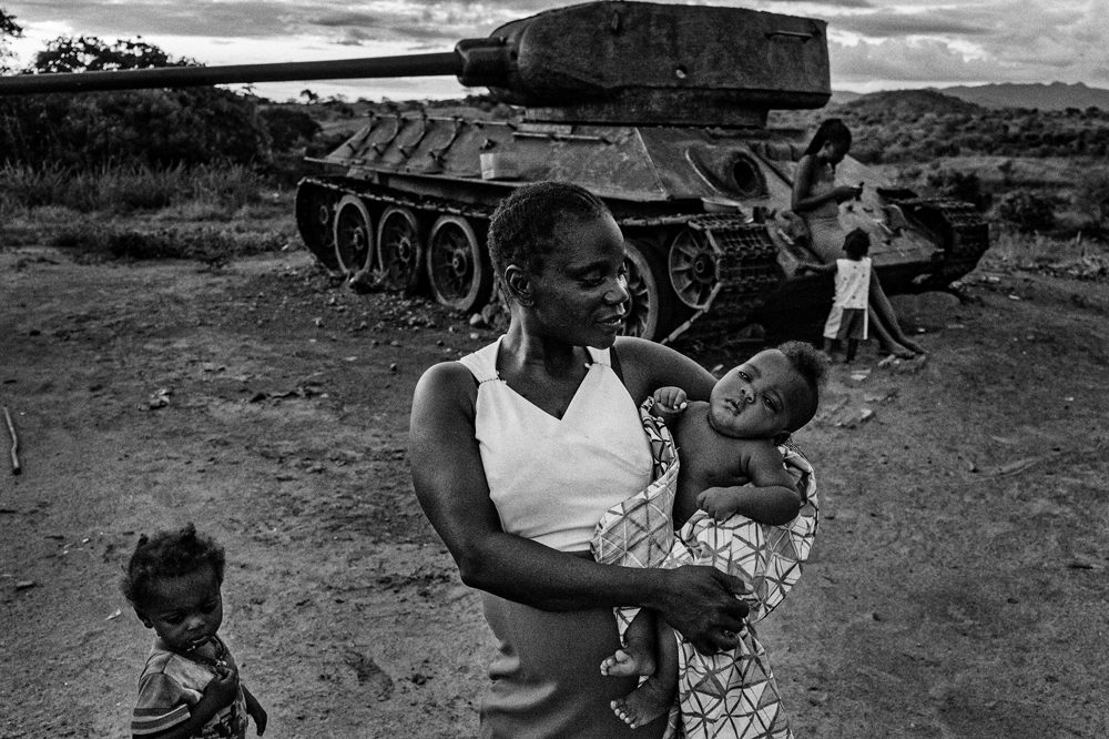A woman holding a baby stnds in front of an abandoned tank that another person is leaning on.
