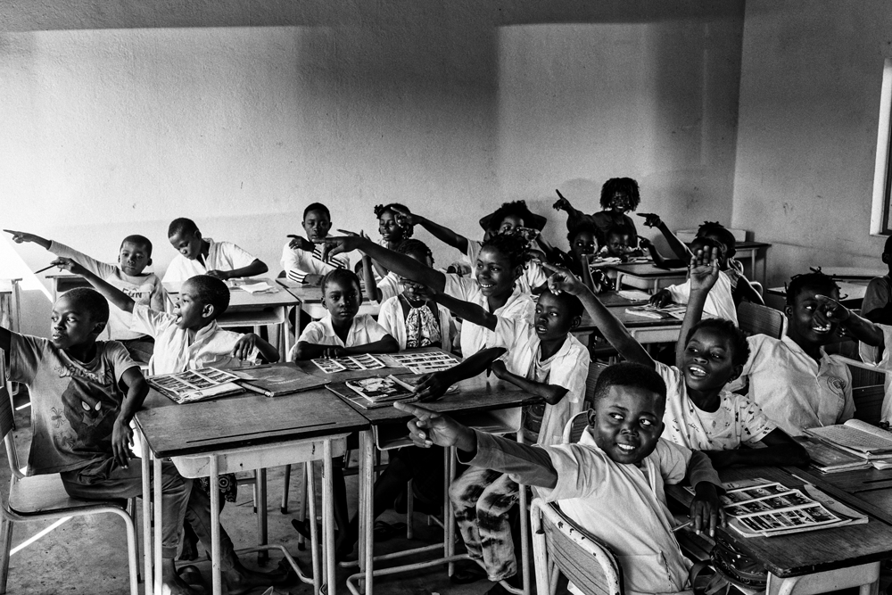 A group of children sitting at desks in a classroom all point to the left with their arms raised.