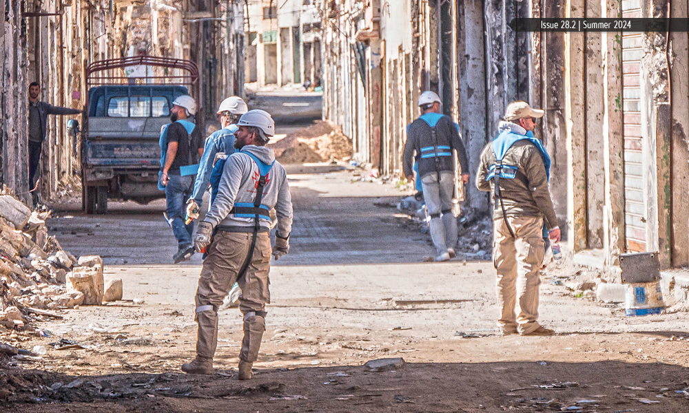 A group of men in personal protective equipment walk down a street and inspect buildings and debris. 