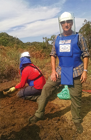 A person wearing a blue protective apron, helmet, and faceshield stands on dirt ground in front of another person kneeling on the ground digging with small tools