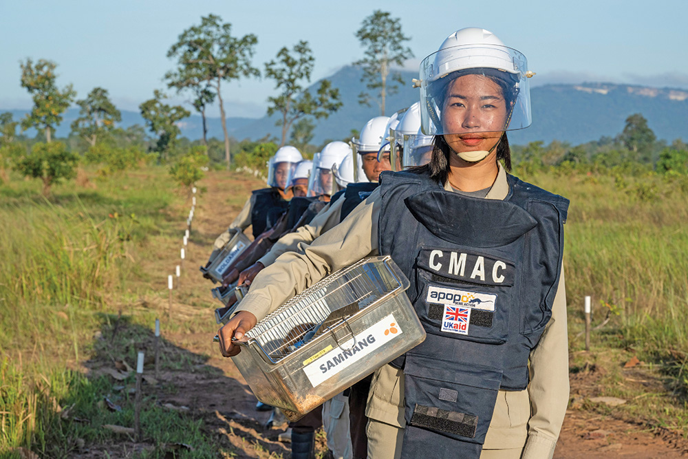 A column of people wearing protective aprons, helmets, and visors walk down a dirt path marked with stakes.