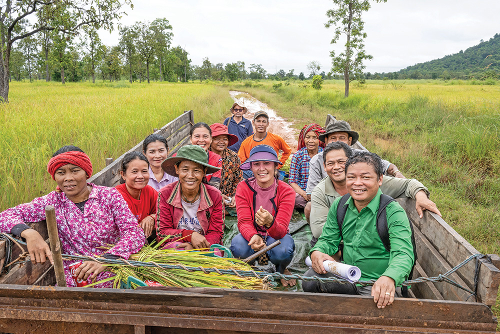 Several people in colorful clothing sit inm the bed of a vehicle going down a path in a ricefield.