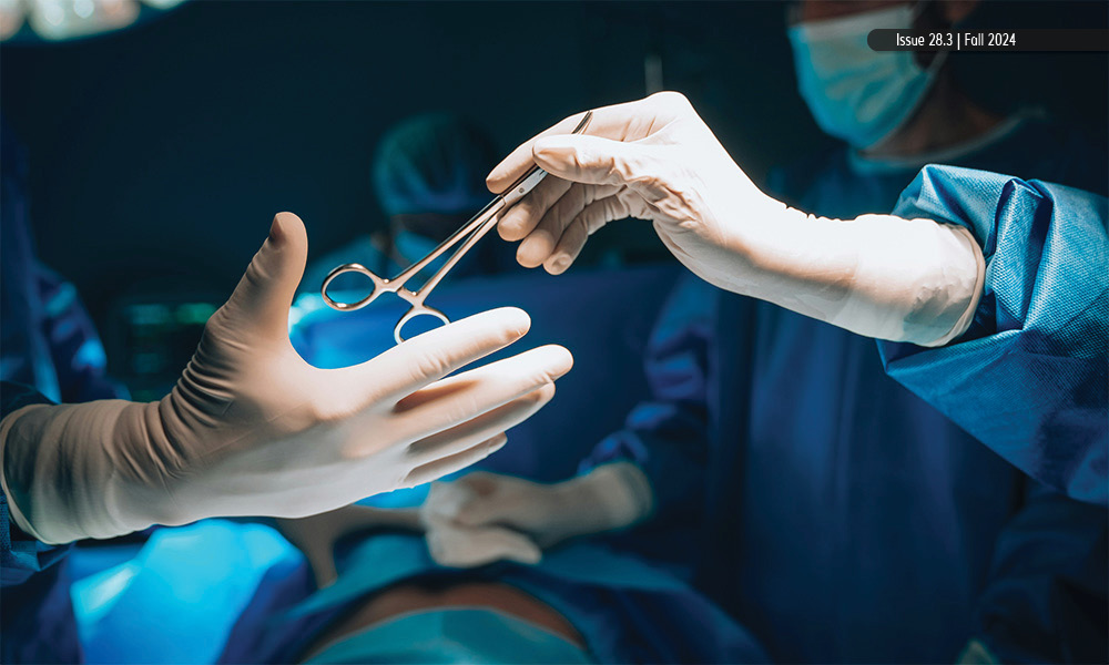 Gloved hands pass off surgical scissors to another set of gloves hands while in an operating room with a surgical team in the background.