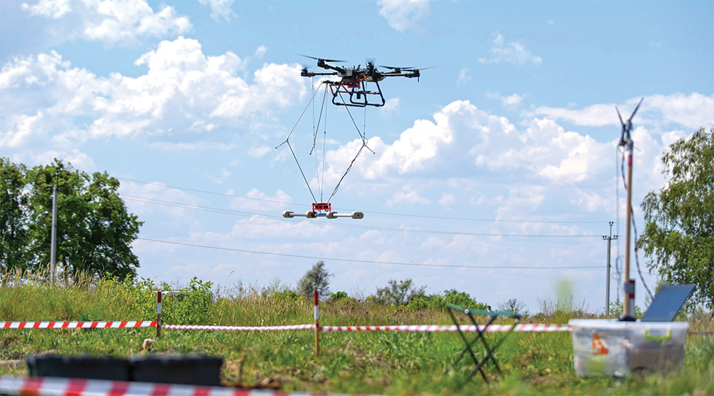 A small machine with propellers hovers in the air with a device shaped like a bar suspended underneath.