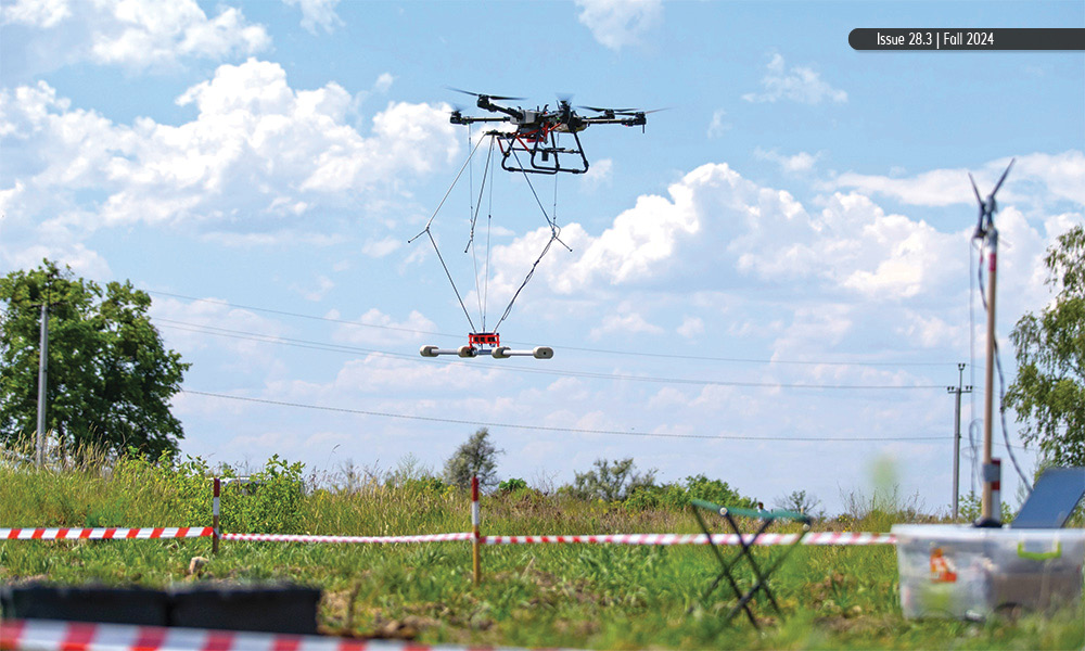 A drone with an attached hanging device flying over a grassy field with caution tape in the background.