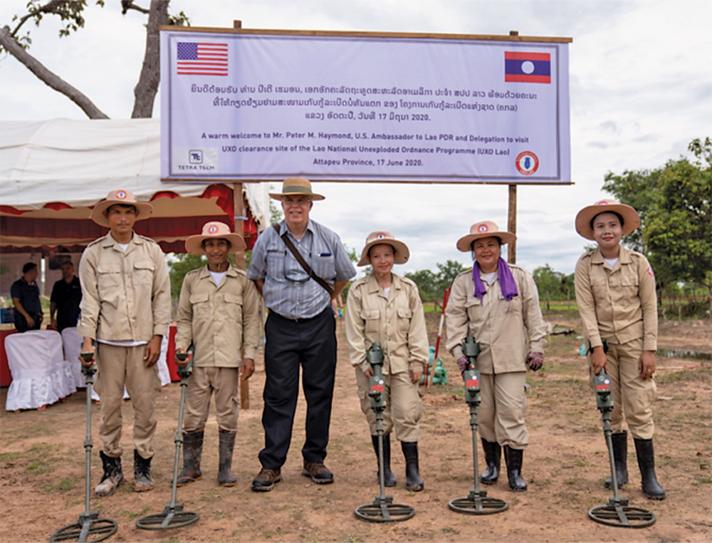 Six people stand in a row, five men and women wear tan fatigues and hold metal detectors, a sixth man stands between them in plain clothes.