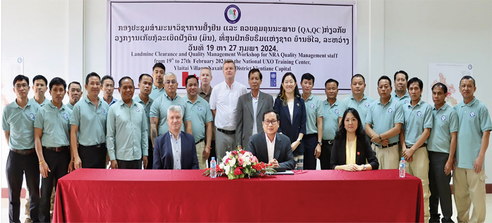 A large group of mostly men wearing light blue shirts stand side-by-side in front of a large banner with Lao script and behind a red table with three seated people in formal attire.