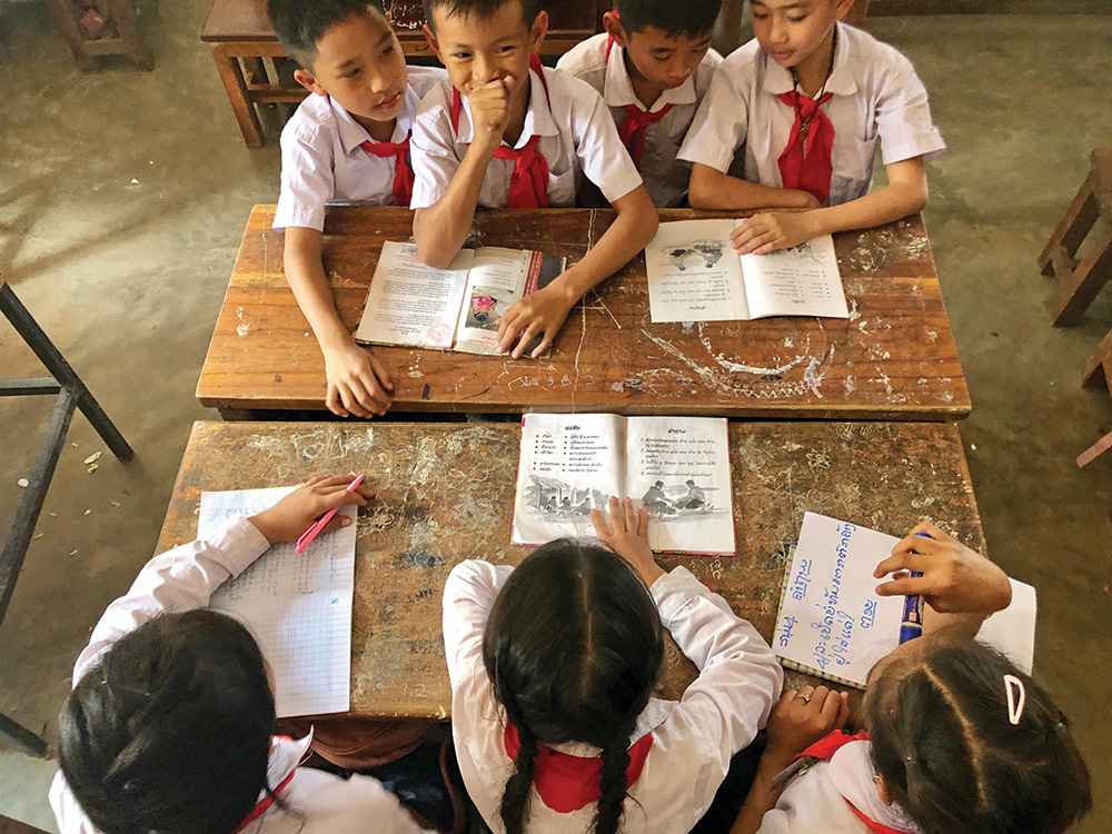 Four young boys side-by-side at a table, one holds his fist over his mouth and three young girls sit side-by-side and look at a booklet with drawing of people with mine risk education messages.