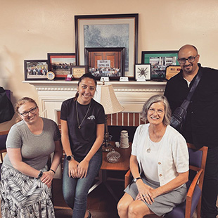 Three women and one smile and pose for a picture by a fireplace.