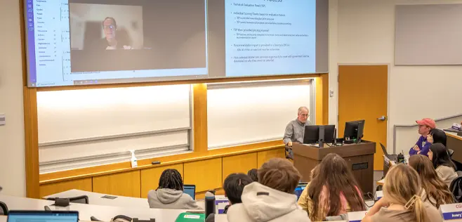 A classroom lecture in progress, featuring a professor speaking to students both in-person and via video conference displayed on a large screen.