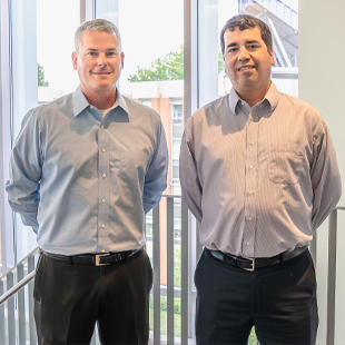 Christian Ryder, left, and Luis Salcedo, right, standing together in Hartman Hall, with large windows and a staircase in the background