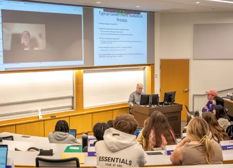 A classroom setting where an instructor is giving a lecture, with students seated at desks and a video conference screen in the background displaying a remote participant.