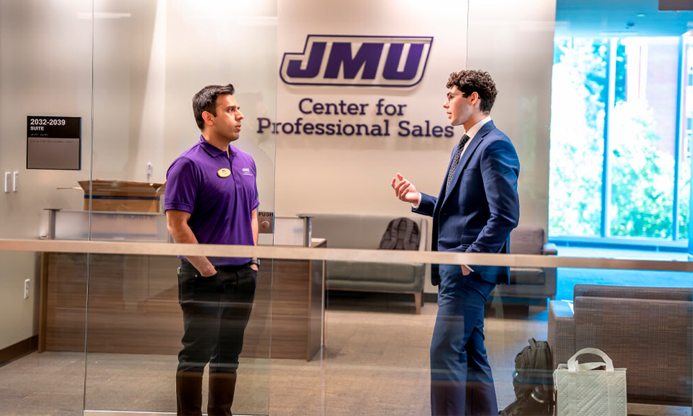 A professor and a student in a suit stand facing each other in front of the glass doors of the Center for Professional Sales in Hartman Hall