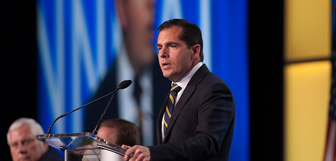 Derek Sprague addressing an audience at a conference, in front of a large screen displaying a blurred background image.