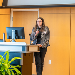 Morgan Tribby presenting at a conference with a microphone in hand, standing beside a podium and a computer screen.