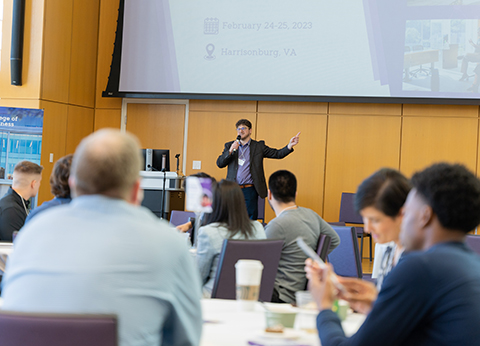 A speaker presenting at a conference in Harrisonburg, VA, with an audience engaged in the background.