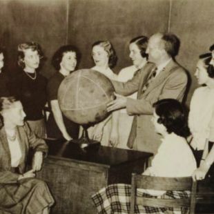 A classroom scene featuring a group of women and Dr. Ikenberry gathered around a globe, engaged in discussion.