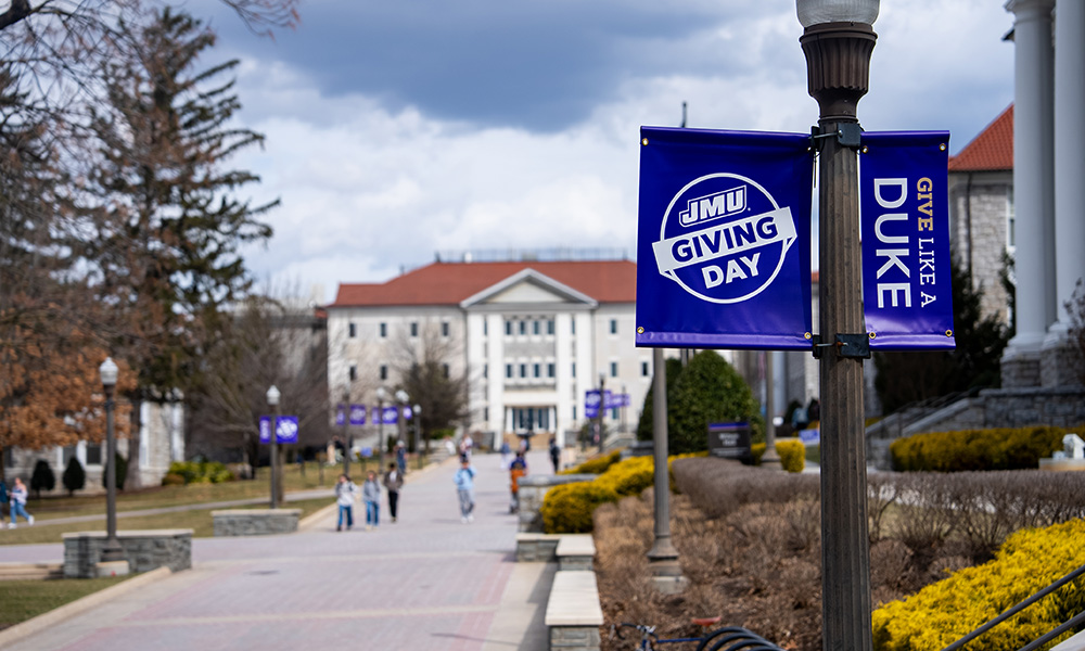 Banner for James Madison University's Giving Day, displaying the slogan "Give Like a Duke" with campus buildings in the background.