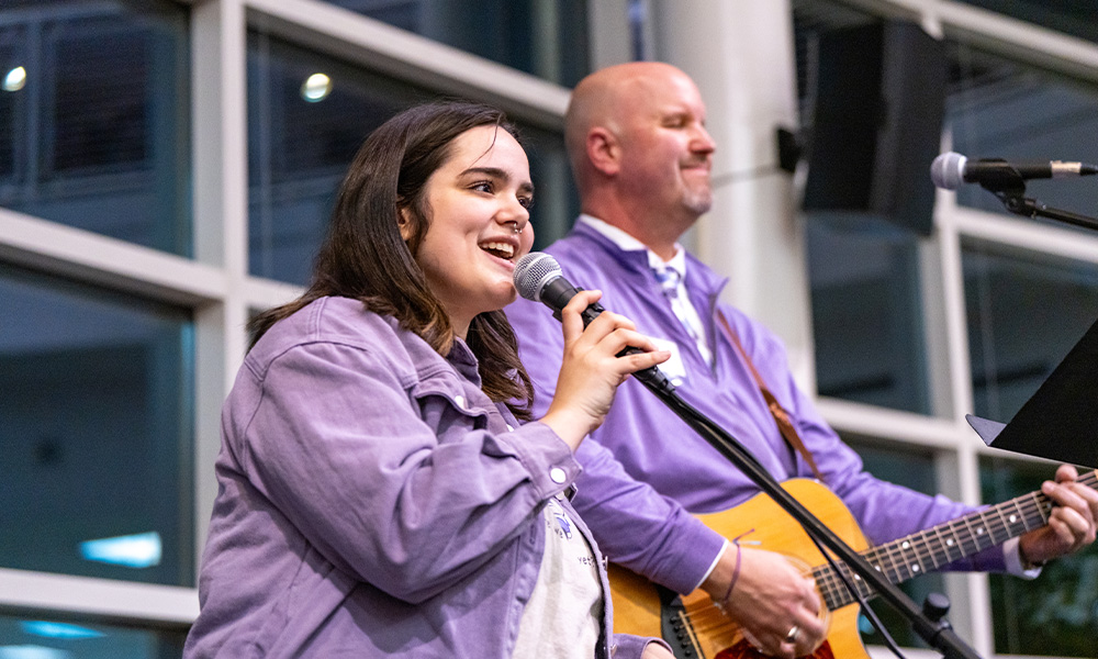 A student sings into a microphone while Tim Miller plays next to her guitar and both appear in a brightly lit venue.