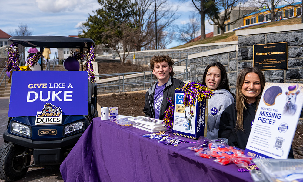 Students who advertise a fundraising campaign at a campus event with a decorated golf car that is decorated with one "Give like a duke" Sign and a decorated table with information materials.