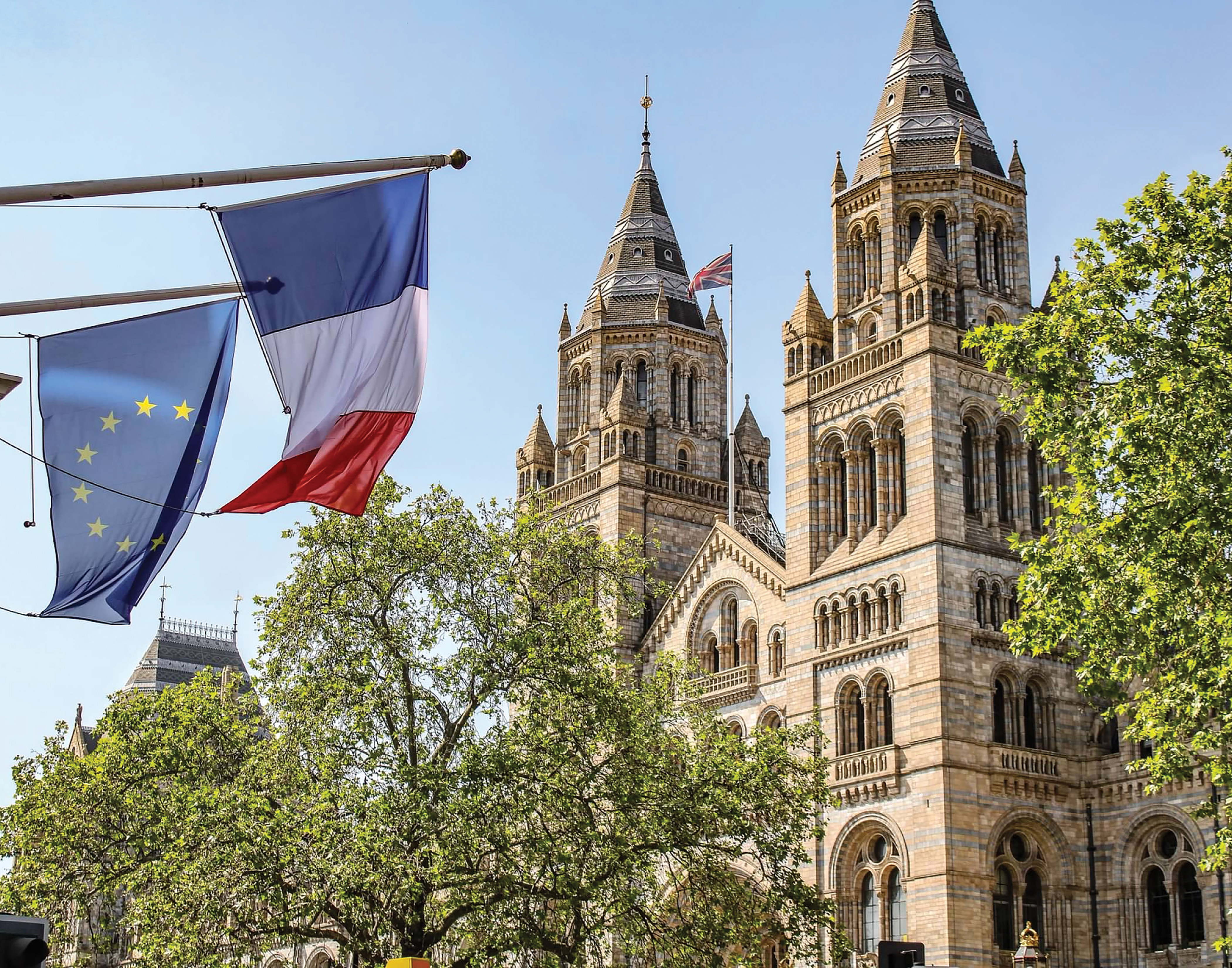 Flags of France and the European Union displayed near a historical building with tall turrets and a clear blue sky.