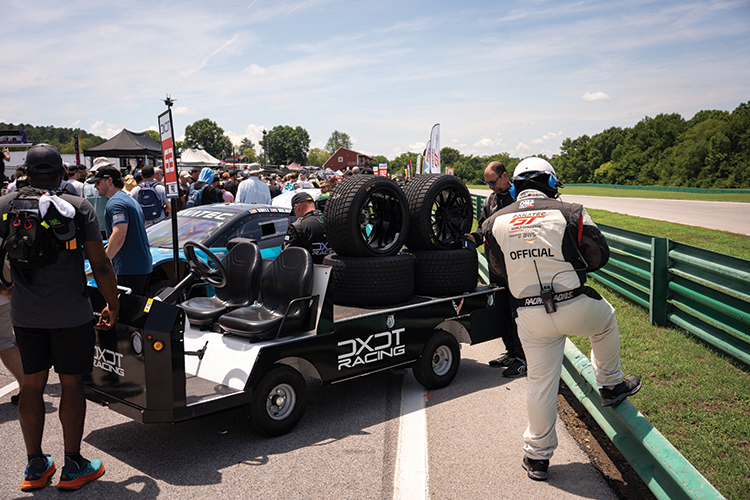 A racing team in a paddock area prepares for an event, featuring crew members and a vehicle equipped with spare tires.