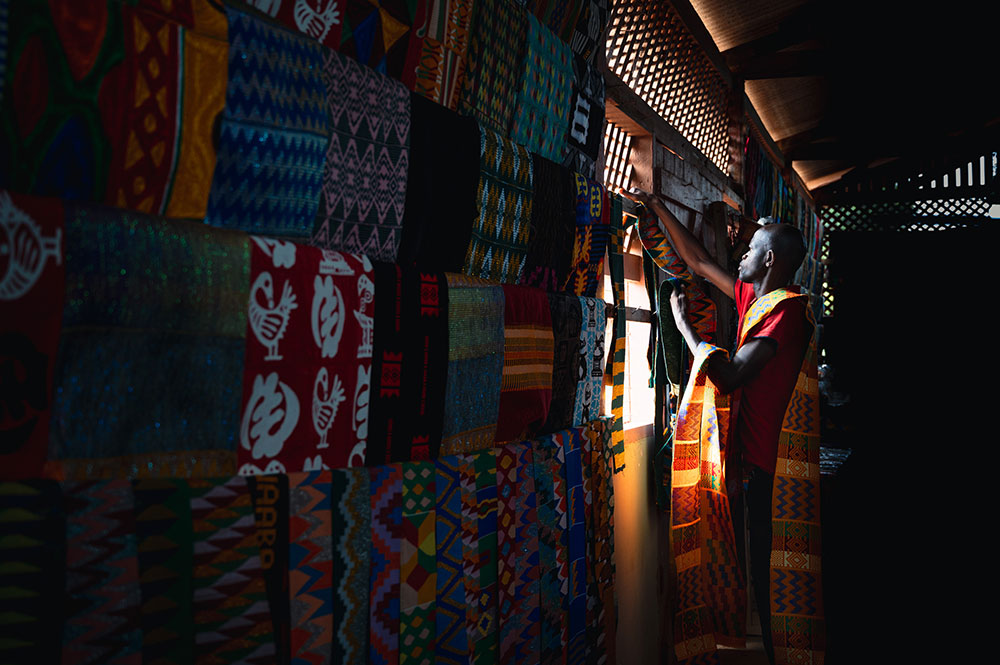 A photo of an individual in a dark room with light coming in a small window. They are holding up pieces of Kente cloth.