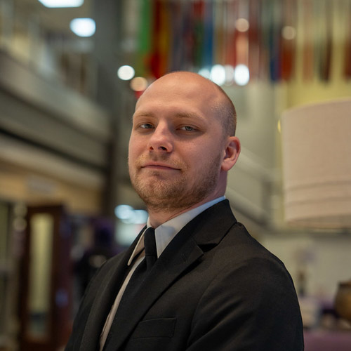 Professional portrait of a man in a suit, exuding confidence with a blurred background featuring colorful flags.