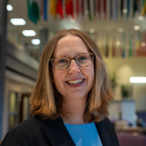 A smiling professional woman with shoulder-length hair and glasses, standing in a well-lit space adorned with flags.