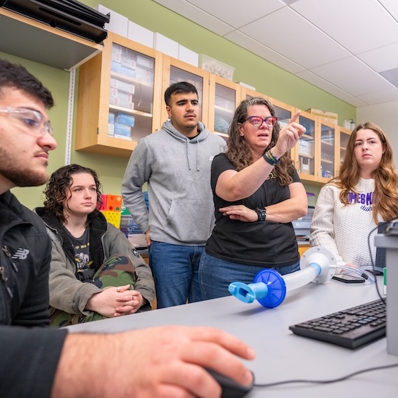 Instructor gestures while students watch a computer demonstration in a science classroom.